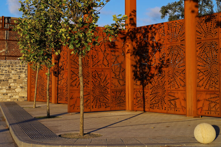 External view of decorative pre-rusted perforated steel fence surrounding the Port Centre in Dublin, Ireland. Trees in the foreground. Sunny day.
